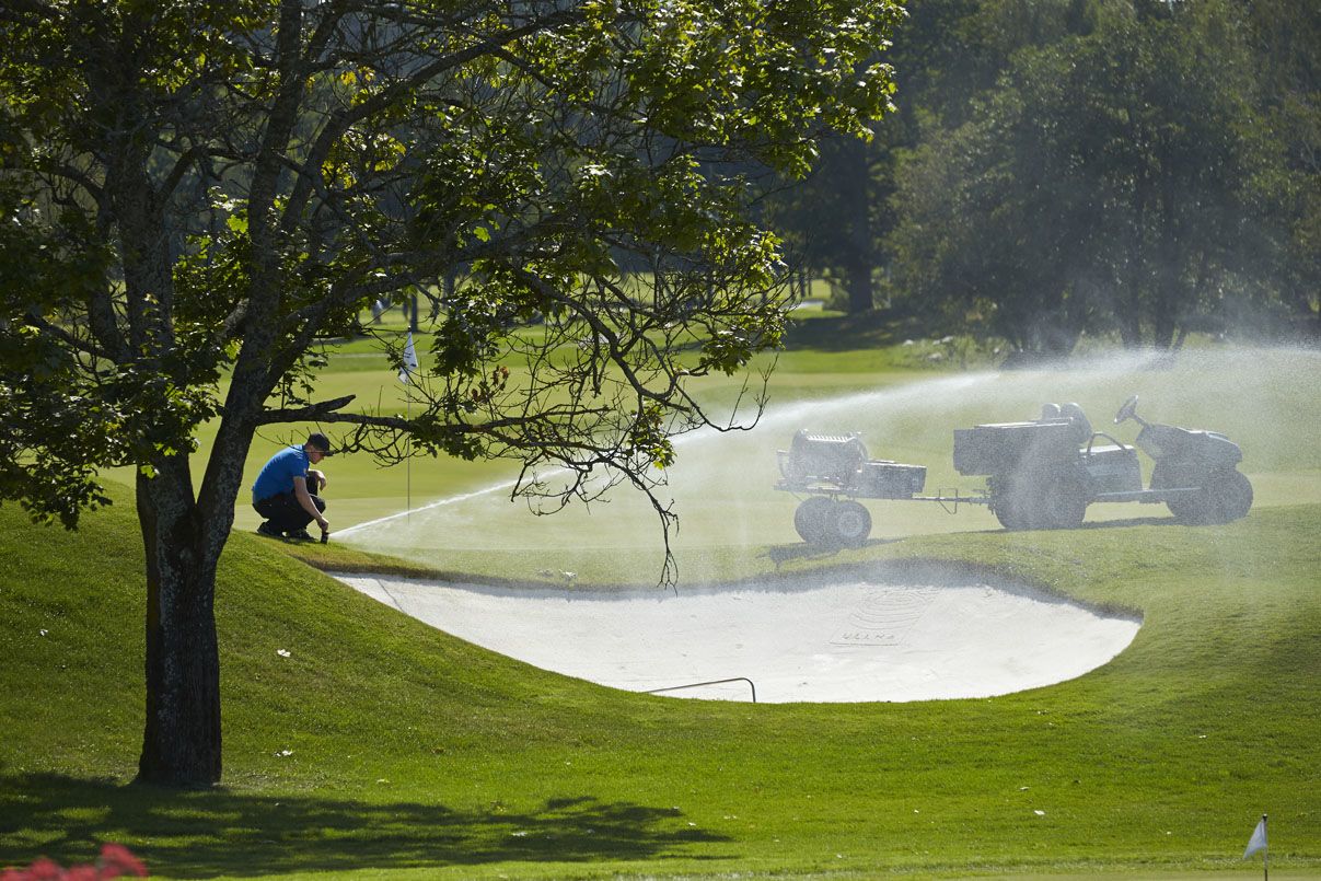 Golf course superintendent at work as sprinkler waters the green.